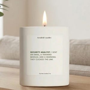 May include: A white, rectangular soy wax candle with a lit flame. The candle has a label with the text "wenfield candles" and a humorous quote about a security analyst. The candle is 9 oz.