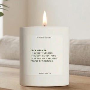 May include: A lit white soy wax candle with the text "wenfield candles" and "DECK OFFICER: I NAVIGATE VESSELS THROUGH CONDITIONS THAT WOULD MAKE MOST PEOPLE RECONSIDER." The candle is 9 oz.