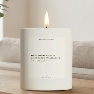 May include: A lit white soy wax candle with the text "MATCHMAKER: I HAVE INTRODUCED 200 COUPLES. 40 MARRIAGES." and "wenfield candles." The candle is 9 oz and sits on a wooden surface.