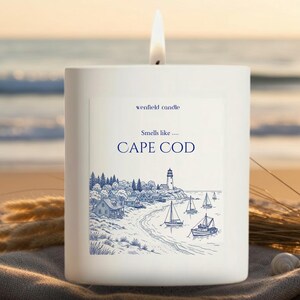May include: A white square candle with a lit wick. The label reads "wenfield candle" and "Smells like... CAPE COD" with a blue illustration of a coastal scene with a lighthouse, boats, and houses. The background is a beach.
