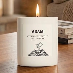 May include: A white cylindrical candle with a lit wick. The label features the text ADAM, CONGRATS ON THE PROMOTION, and a black and white line drawing of a pile of paper money with one bill floating above the stack.