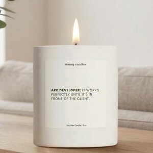 May include: A white soy wax candle with a lit flame. The candle has a rectangular shape and features the text "sunny candles" and a humorous quote about app developers. The candle is 9 oz.