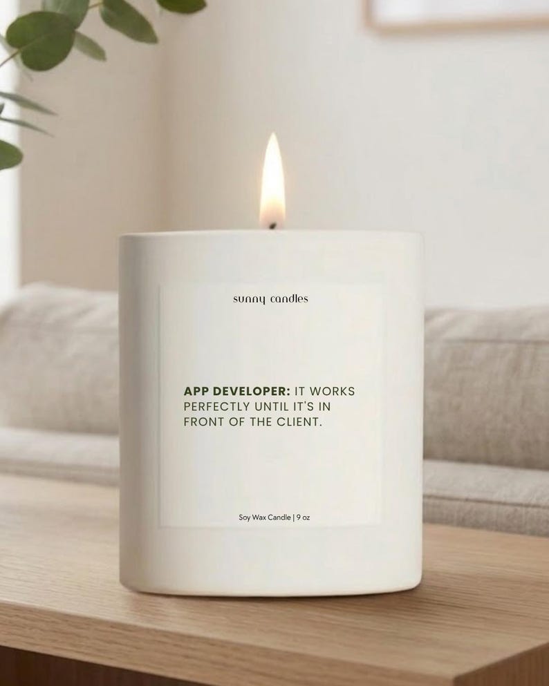 May include: A white soy wax candle with a lit flame. The candle has a rectangular shape and features the text "sunny candles" and a humorous quote about app developers. The candle is 9 oz.