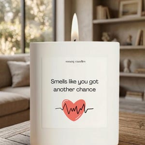 May include: A white, rectangular candle with a lit wick. The candle has a label that reads "Smells like you got another chance" with a red heart and heartbeat graphic. The text "sunny candles" is also on the label.