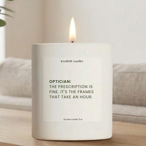 May include: A lit white soy wax candle with the text "OPTICIAN: THE PRESCRIPTION IS FINE. IT'S THE FRAMES THAT TAKE AN HOUR." The candle is 9 oz and is on a wooden surface. The brand name "wenfield candles" is also visible.