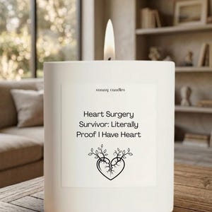 May include: A white square candle with a lit wick. The candle has a label with the text "Heart Surgery Survivor: Literally Proof I Have Heart" and a graphic of a broken heart with leaves. The brand name "sunny candles" is also visible.