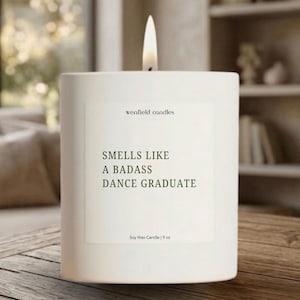 May include: A white square candle with a lit wick. The candle has a label that reads "wenfield candles" and "Smells Like A Badass Dance Graduate." It is a soy wax candle, 9 oz.