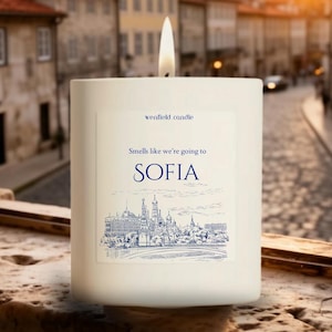 May include: A lit white candle with a label that reads "wenfield candle" and "Smells like we're going to SOFIA." The label features a blue illustration of a city skyline. The candle is in front of a blurred city background.