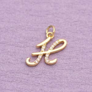 Pode incluir: Um pingente de metal em tom dourado com o formato da letra H em cursivo, apresentando um acabamento polido e uma série de pequenas pedras claras e brilhantes incrustadas na superfície frontal, com um pequeno anel circular na parte superior.