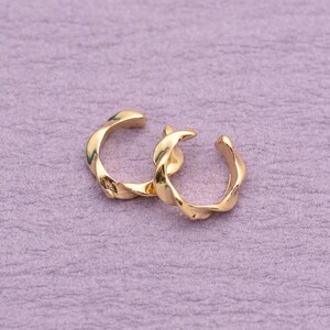 Puede incluir: Un par de pendientes tipo ear cuff dorados con un diseño trenzado similar a una cuerda. Cada pieza es circular con una abertura, mostrando una superficie metálica pulida y reflectante que capta la luz, creando un aspecto fluido y texturizado.