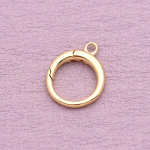 May include: A circular gold-toned spring ring clasp featuring a smooth, polished finish and a small integrated loop at the top. The mechanism includes a spring-loaded gate that opens and closes to secure jewelry components or charms in place.