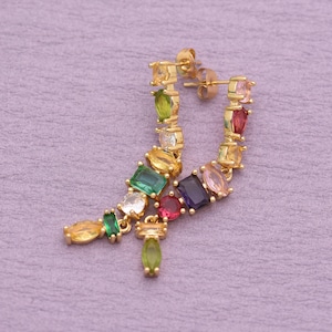 May include: A pair of gold-tone earrings featuring a linear arrangement of multi-colored gemstones in various shapes, including marquise, emerald, and round cuts. The stones display hues of green, yellow, pink, purple, and clear white.