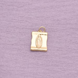 May include: A gold-toned rectangular pendant featuring a raised, embossed depiction of the Virgin of Guadalupe centered on the surface. The pendant has a smooth, polished finish and includes a small circular jump ring attached to the top edge.