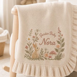 May include: A cream-colored knit blanket featuring a ruffled edge and an embroidered design of a rabbit, mushrooms, and wildflowers. The text "Garden Baby Nora" is stitched in the center of the floral wreath in a brown script font.