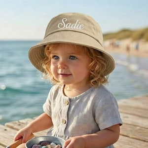May include: A tan bucket hat with the name "Sadie" in white script. The hat is worn by a young child, who is holding a small metal bucket filled with colorful stones. The child is wearing a light gray dress.