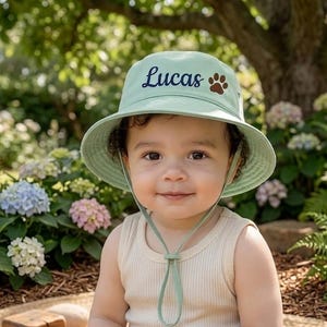 May include: A light green bucket hat with the name "Lucas" in cursive and a brown paw print. The hat has a chin strap and a wide brim. The child is wearing a beige tank top.