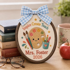 Puede incluir: Un bastidor de bordado ovalado con un lazo de cuadros vichy azul claro en la parte superior. La tela muestra artículos de arte bordados, incluyendo una paleta, pinceles y lápices, con el texto "Inspire Create Teach", "Mrs. Foster" y "2026".