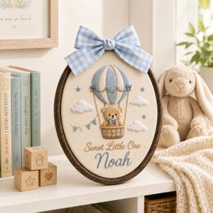 May include: An oval embroidery hoop featuring a light blue and white gingham bow at the top. The fabric displays a stitched hot air balloon carrying a brown teddy bear, white clouds, blue stars, and the text Sweet Little One Noah in blue thread.