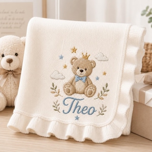 Puede incluir: Una manta de punto color crema para bebé con borde de volantes y un diseño bordado de un oso de peluche marrón con corona dorada y pajarita azul. El diseño incluye nubes, estrellas, ramas verdes y el nombre Theo bordado en color azul.