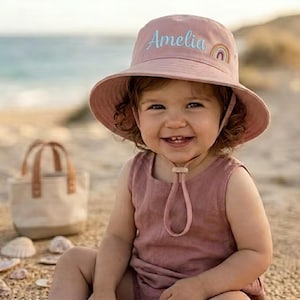 May include: A pink sun hat with the name "Amelia" embroidered in light blue, and a rainbow design. The hat has a wide brim and a chin strap. The child is wearing a pink dress and is smiling. A small tote bag is in the background.