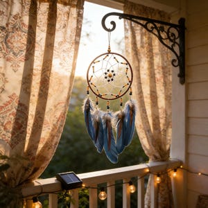 Op de afbeelding: Een handgemaakte dromenvanger met een bruin cirkelvormig frame, een ingewikkeld webontwerp en blauwe veren. De dromenvanger hangt aan een decoratieve zwarte metalen beugel op een veranda, met lichtslingers en gordijnen met patronen op de achtergrond.