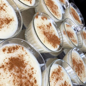 May include: A close-up shot of several glass bowls filled with a creamy white dessert, each dusted with a generous sprinkle of cinnamon. The desserts are arranged in rows, suggesting a batch of homemade treats.