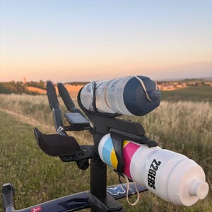 May include: A triathlon bicycle cockpit featuring two water bottles mounted on the aero bars. The top bottle is translucent with a dark blue cap, while the bottom bottle is white with blue, yellow, and pink stripes and the text 226ERS.