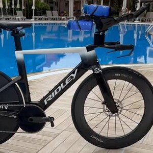 May include: A black and white Ridley triathlon bicycle with aero bars and disc wheels. The bike has a water bottle cage and is positioned near a pool. The rear wheel has "DT Swiss" and "1100" printed on it.