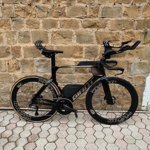 May include: A black Giant road bike with aerodynamic handlebars and wheels. The bike has a black frame with gold accents and is positioned in front of a brick wall. The wheels have the number "60" on them.