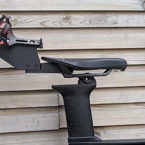 Puede incluir: Sillín de bicicleta negro con un portabidón adjunto. El sillín está montado en un poste y un cuadro negros. El portabidón es negro y rojo. Sobre un fondo de pared de madera.