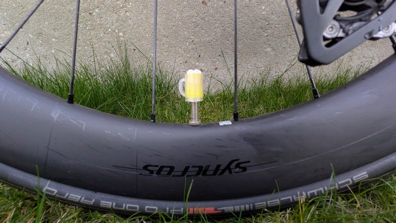 May include: A bicycle wheel with a yellow beer-shaped valve cap. The wheel is black with the word "SYNEROS" printed on the side. The background is green grass.