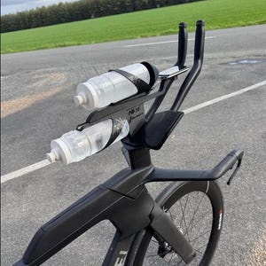 May include: Black triathlon bike with two water bottles mounted on the handlebars. The bike features aero bars and a streamlined design. The background shows a road, green field, and trees.