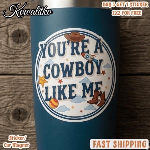 Op de afbeelding: Een donkerblauwe beker met een ronde sticker met de tekst YOU'RE A COWBOY LIKE ME. Het ontwerp bevat illustraties van een bruine cowboyhoed, een raket, een gele ster, een gestreepte bal en bruine cowboylaarzen tussen kleine sterren en wolken.