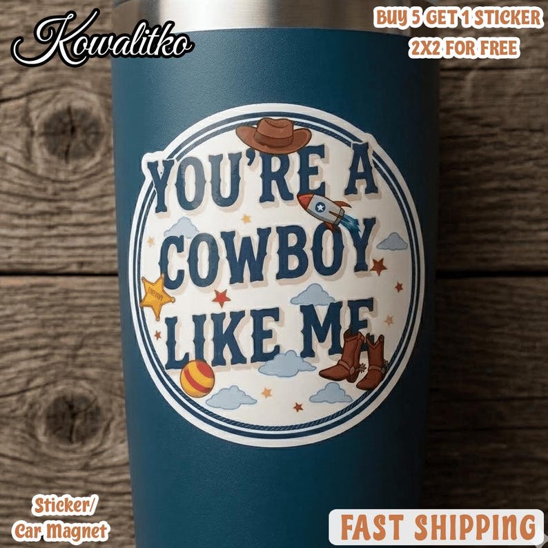 Op de afbeelding: Een donkerblauwe beker met een ronde sticker met de tekst YOU'RE A COWBOY LIKE ME. Het ontwerp bevat illustraties van een bruine cowboyhoed, een raket, een gele ster, een gestreepte bal en bruine cowboylaarzen tussen kleine sterren en wolken.