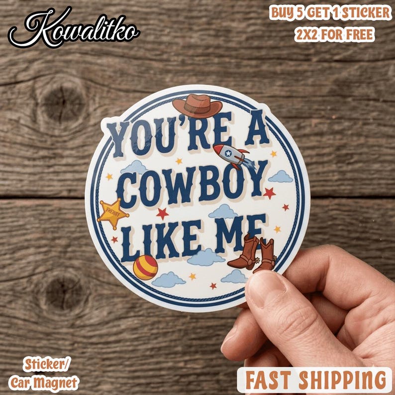 Op de afbeelding: Een ronde sticker met de tekst YOU'RE A COWBOY LIKE ME in donkerblauwe letters. Het ontwerp bevat een bruine cowboyhoed, een raket, een gele sheriffster, een gestreepte bal en bruine cowboylaarzen, allemaal omringd door kleine blauwe wolken en rode sterren.