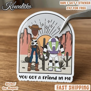 Puede incluir: Una pegatina troquelada con una ilustraci&oacute;n de dibujos animados de un vaquero y un guardi&aacute;n espacial en un paisaje des&eacute;rtico con cactus y un sol. El texto "YOU GOT A FRIEND IN ME" est&aacute; impreso en negro en la parte inferior de la pegatina.