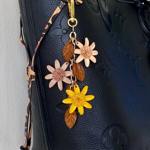 K&ouml;nnte beinhalten: Schwarze Tragetasche mit gepr&auml;gtem Blumenmuster, mit einem dekorativen Anh&auml;nger. Der Anh&auml;nger enth&auml;lt einen goldfarbenen Verschluss, braune Bl&auml;tter und rosa und gelbe Blumenakzente. Die Tasche hat einen Leopardenmuster-Gurt.