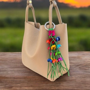 May include: A beige leather handbag with a colourful flower charm attached to the handle. The charm features a cluster of small, brightly coloured flowers with green stems.