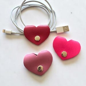 May include: Three heart-shaped cable tidies in shades of red and pink, each with a gold snap closure. A grey cable is looped through a red tidy. The tidies are designed to manage and secure cords.
