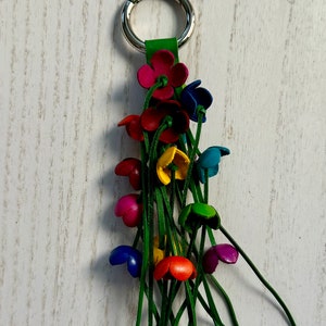 K&ouml;nnte beinhalten: Ein bunter Leder-Schl&uuml;sselanh&auml;nger mit einem silbernen Ring und einem Quasten aus gr&uuml;nen Lederstr&auml;ngen. Der Schl&uuml;sselanh&auml;nger zeigt mehrere kleine Blumen in verschiedenen Farben, darunter Rot, Pink, Blau, Gelb und Gr&uuml;n.