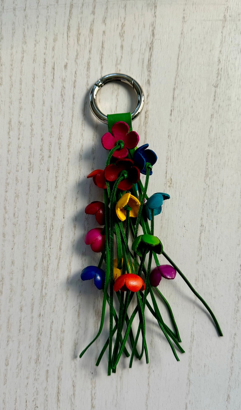 K&ouml;nnte beinhalten: Ein bunter Leder-Schl&uuml;sselanh&auml;nger mit einem silbernen Ring und einem Quasten aus gr&uuml;nen Lederstr&auml;ngen. Der Schl&uuml;sselanh&auml;nger zeigt mehrere kleine Blumen in verschiedenen Farben, darunter Rot, Pink, Blau, Gelb und Gr&uuml;n.