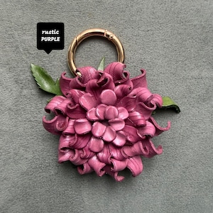 Op de afbeelding: Een handgemaakte, rustieke paarse leren bloem sleutelhanger met een goudkleurige sluiting. De bloem heeft gedetailleerde bloemblaadjes en groene bladeren. De tekst "rustic PURPLE" staat in een zwarte doos.
