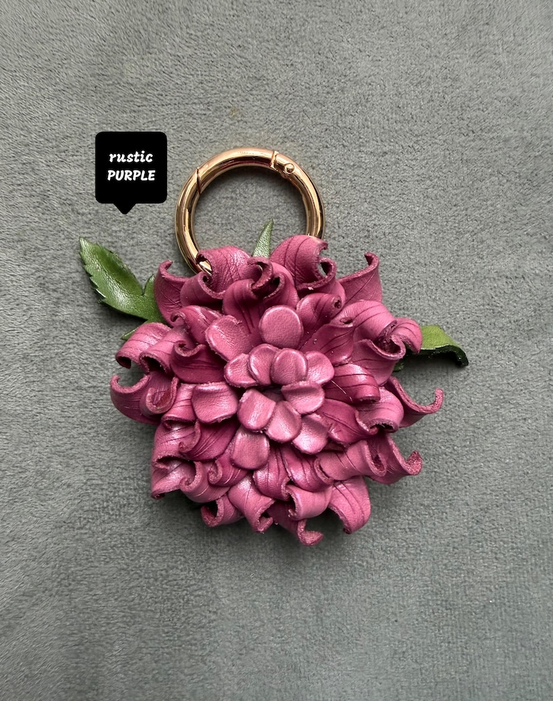 Op de afbeelding: Een handgemaakte, rustieke paarse leren bloem sleutelhanger met een goudkleurige sluiting. De bloem heeft gedetailleerde bloemblaadjes en groene bladeren. De tekst "rustic PURPLE" staat in een zwarte doos.