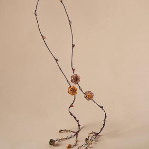 May include: A long necklace featuring a thin dark brown cord adorned with small, multi-colored flower-shaped beads in shades of orange, brown, and green. The design includes scattered small beads along the cord and dangling floral accents at the ends.