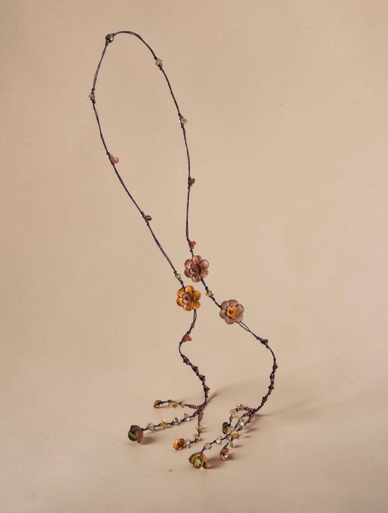 May include: A long necklace featuring a thin dark brown cord adorned with small, multi-colored flower-shaped beads in shades of orange, brown, and green. The design includes scattered small beads along the cord and dangling floral accents at the ends.