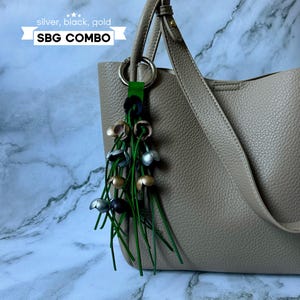 K&ouml;nnte beinhalten: Eine beige Tote Bag mit einem silbernen, schwarzen und goldenen Quasten-Schl&uuml;sselanh&auml;nger. Der Schl&uuml;sselanh&auml;nger hat ein gr&uuml;nes Lederband und der Text "SBG COMBO" ist auf einem wei&szlig;en Banner gedruckt.