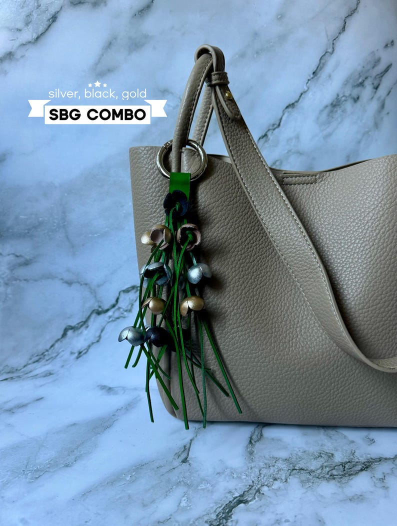 K&ouml;nnte beinhalten: Eine beige Tote Bag mit einem silbernen, schwarzen und goldenen Quasten-Schl&uuml;sselanh&auml;nger. Der Schl&uuml;sselanh&auml;nger hat ein gr&uuml;nes Lederband und der Text "SBG COMBO" ist auf einem wei&szlig;en Banner gedruckt.