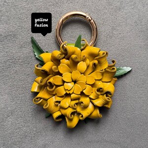 Op de afbeelding: Een handgemaakte gele leren bloem met een goudkleurige metalen sluiting. De bloem heeft groene bladeren en de tekst "yellow fusion" in een zwarte doos. Een helder, vrolijk accessoire.
