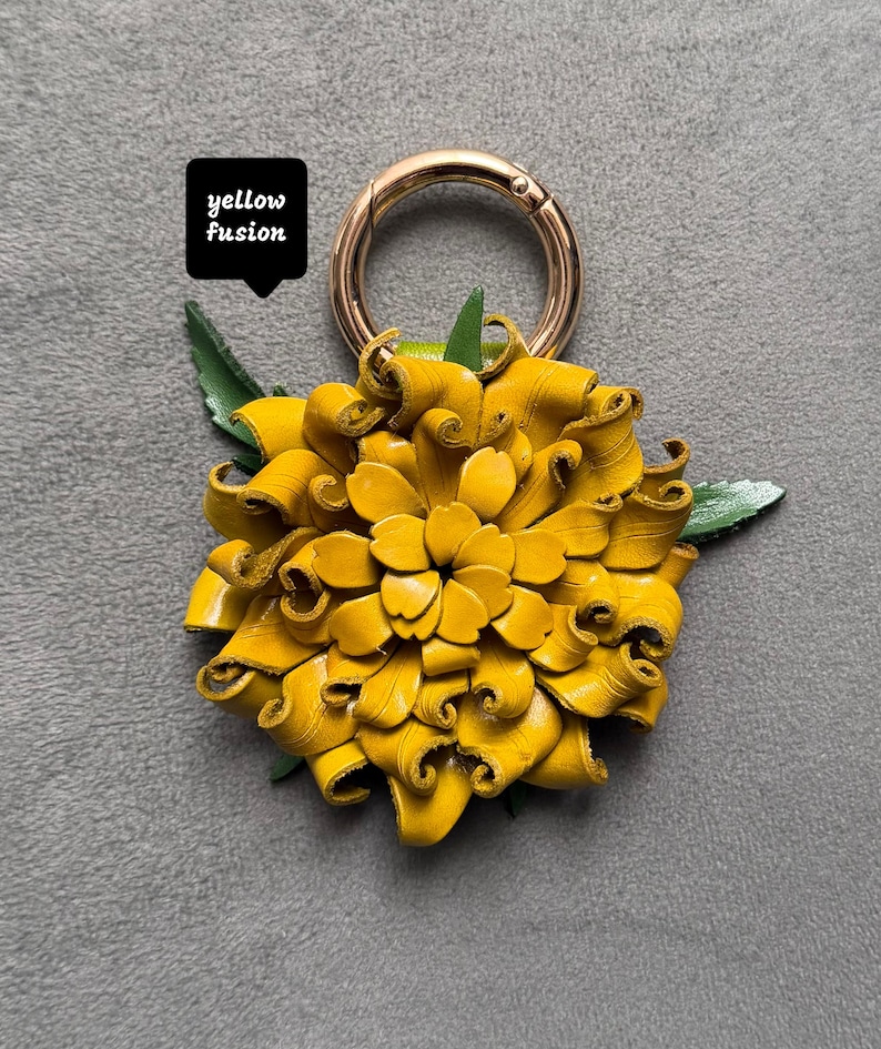 Op de afbeelding: Een handgemaakte gele leren bloem met een goudkleurige metalen sluiting. De bloem heeft groene bladeren en de tekst "yellow fusion" in een zwarte doos. Een helder, vrolijk accessoire.