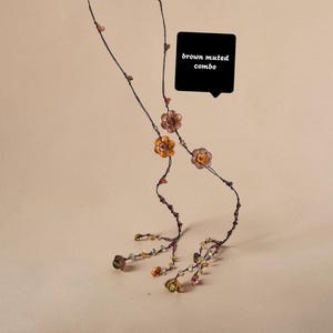 May include: A long, delicate necklace featuring thin dark brown cord strands adorned with small, metallic-toned flower charms and scattered beads in earthy hues. A black speech bubble graphic contains the white text brown muted combo.